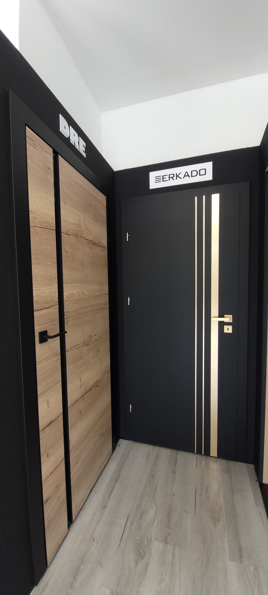 Ekspozycja dwóch modeli drzwi wewnętrznych: drewniane drzwi z poziomym usłojeniem i czarne drzwi z trzema złotymi paskami, oba z czarnymi ościeżnicami, prezentowane na jasnej podłodze w salonie...