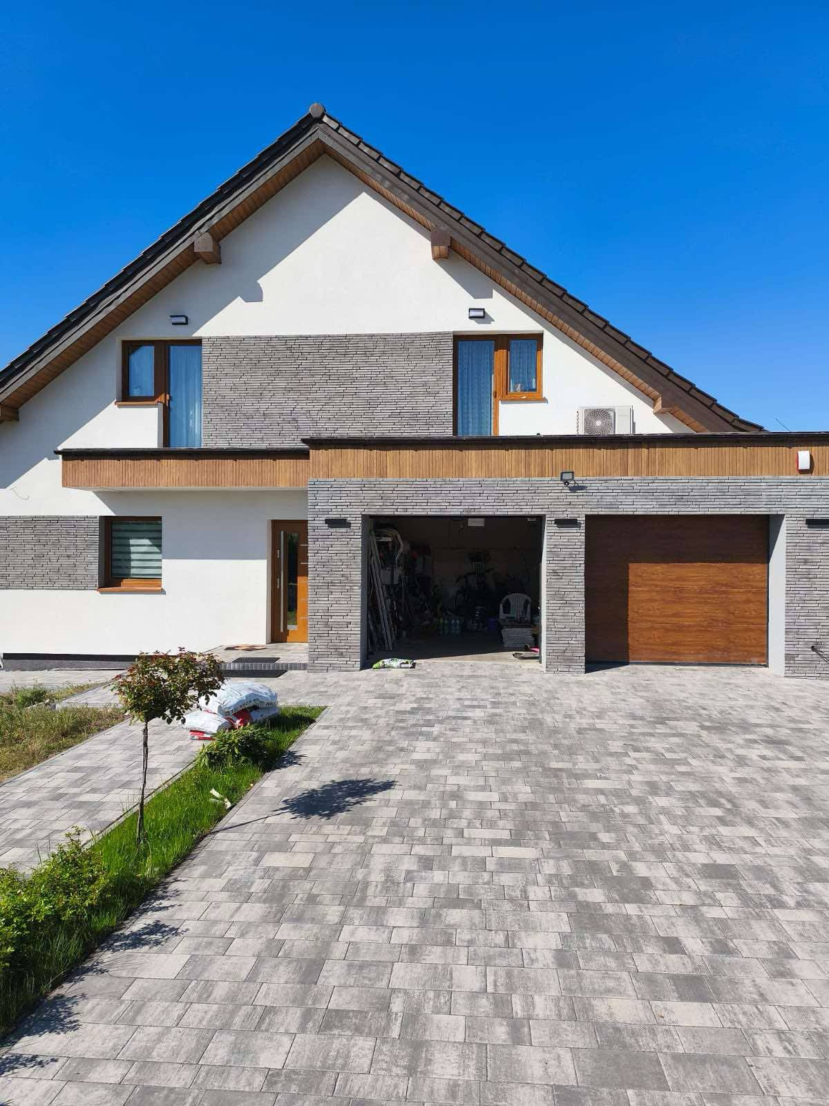 Dwukondygnacyjny dom z szarą elewacją imitującą kamień, białymi ścianami i drewnianymi akcentami, garaż z dwiema bramami i podjazd z kostki brukowej w słoneczny dzień.