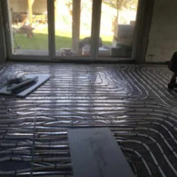 Rozłożone rury ogrzewania podłogowego na folii izolacyjnej w surowym wnętrzu budynku, widoczne narzędzia i płyty izolacyjne, światło słoneczne wpadające przez okno.