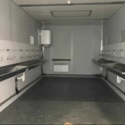 Wnętrze kontenera sanitarnego z rzędami umywalek ze stali nierdzewnej po obu stronach, grzejnikiem ściennym i bojlerem, widoczne instalacje hydrauliczne.