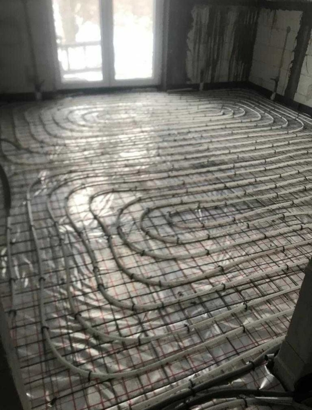 Instalacja ogrzewania podłogowego w surowym pomieszczeniu, rury ułożone na folii i siatce zbrojeniowej, widoczne ściany w stanie deweloperskim i okno.