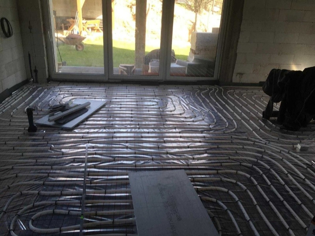 Rozłożone rury ogrzewania podłogowego na folii izolacyjnej w surowym wnętrzu budynku, widoczne narzędzia i płyty izolacyjne, światło słoneczne wpadające przez okno.