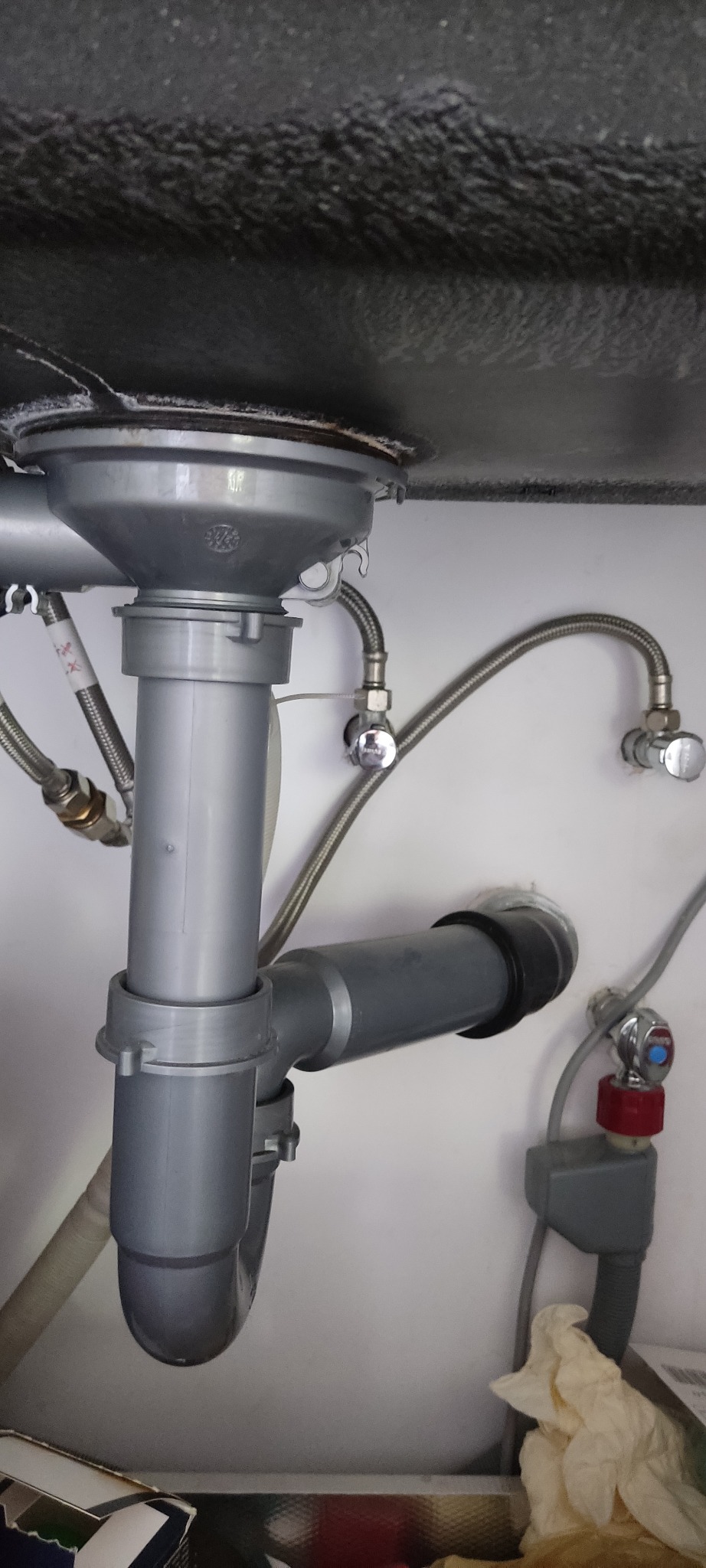 Szare rury odpływowe pod zlewem kuchennym, widoczne wężyki doprowadzające wodę i zawory. Ujęcie od dołu, widoczna powierzchnia zlewu.