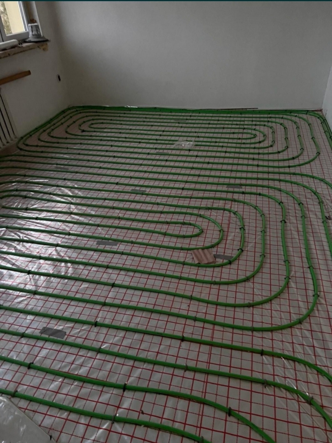 Zielone rurki ogrzewania podłogowego ułożone na folii z czerwoną kratką, widok z góry na instalację w surowym stanie.