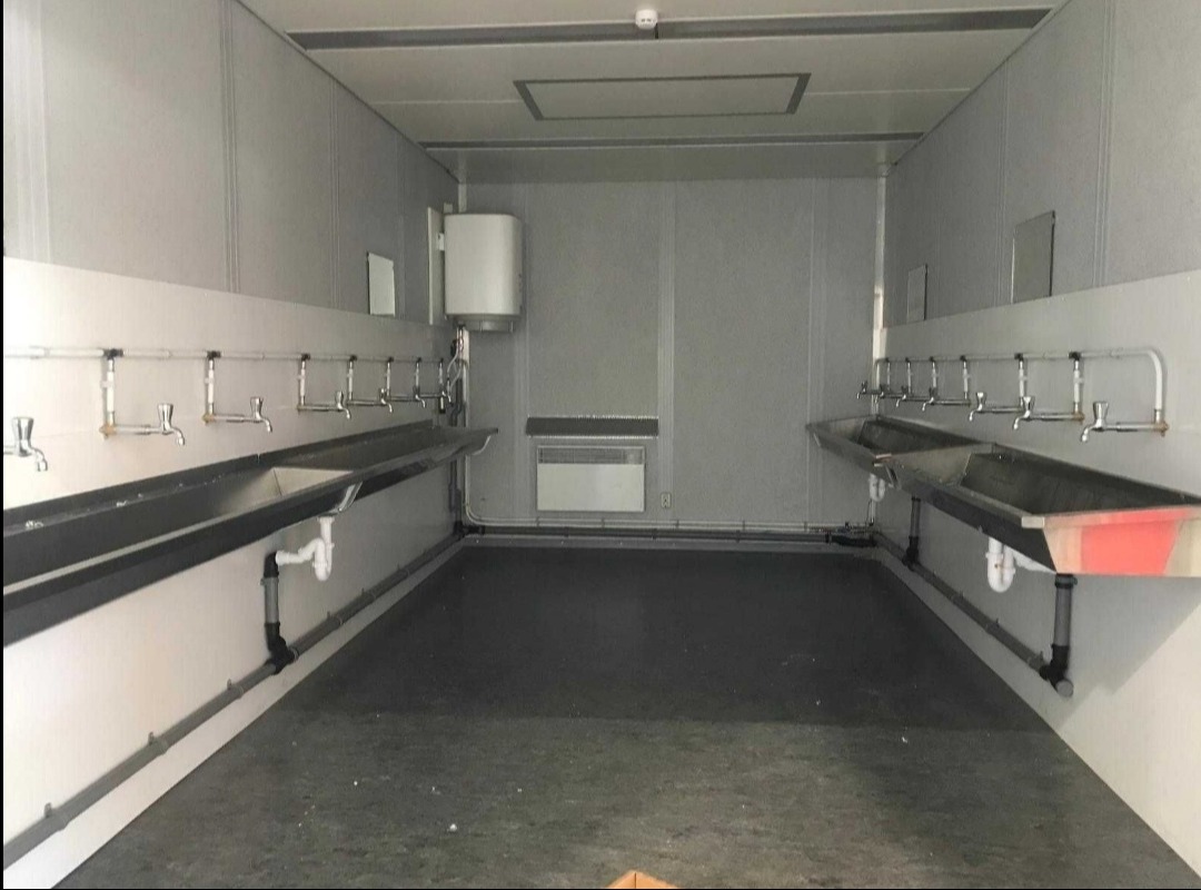 Wnętrze kontenera sanitarnego z rzędami umywalek ze stali nierdzewnej po obu stronach, grzejnikiem ściennym i bojlerem, widoczne instalacje hydrauliczne.