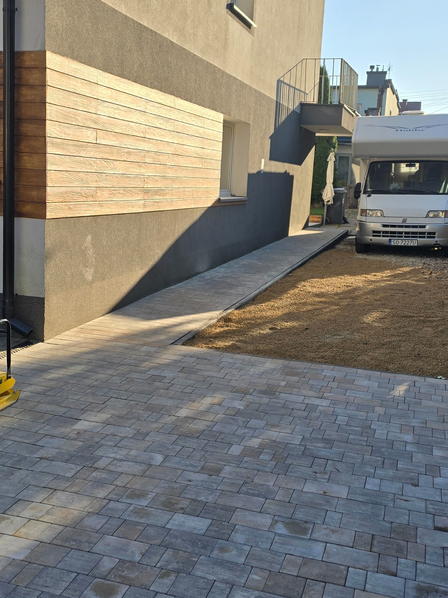 Ułożona kostka brukowa w szarym odcieniu, prowadząca do budynku z drewnianą elewacją i podjazdem z kruszywa. Widoczna zagęszczarka w rogu kadru.