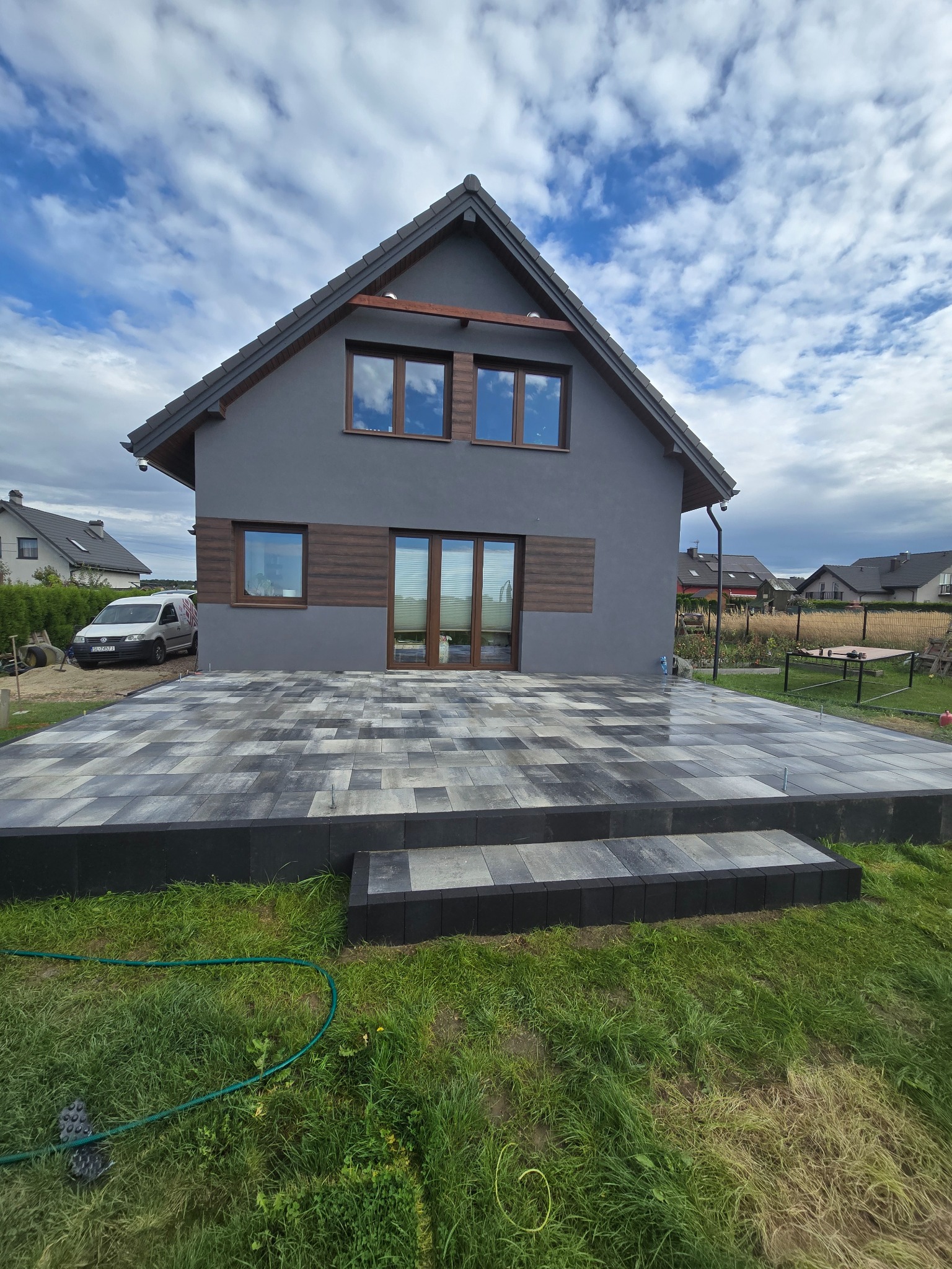 Nowoczesny taras z kostki brukowej w odcieniach szarości, z niskimi schodami, przylegający do domu z szarą elewacją i drewnianymi akcentami. Widok na błękitne niebo z chmurami.