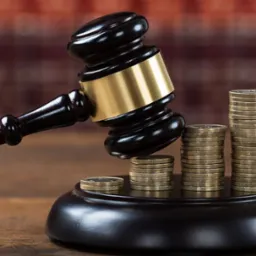 Drewniany młotek sędziowski na podstawie, obok niego trzy stosy monet o różnej wysokości, na drewnianym blacie i ceglanym tle.