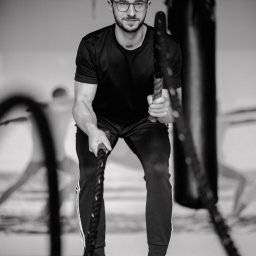 Trener personalny Kraków - Michał Zając - Czarno-białe zdjęcie mężczyzny w okularach, w sportowym stroju, trzymającego grube liny do ćwiczeń w siłowni, z workiem treningowym w tle.