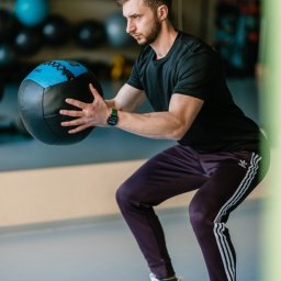 Trener personalny Kraków - Michał Zając - Młody mężczyzna w czarnej koszulce i fioletowych spodniach Adidas wykonuje przysiad z niebiesko-czarną piłką lekarską w siłowni, patrząc w prawo.
