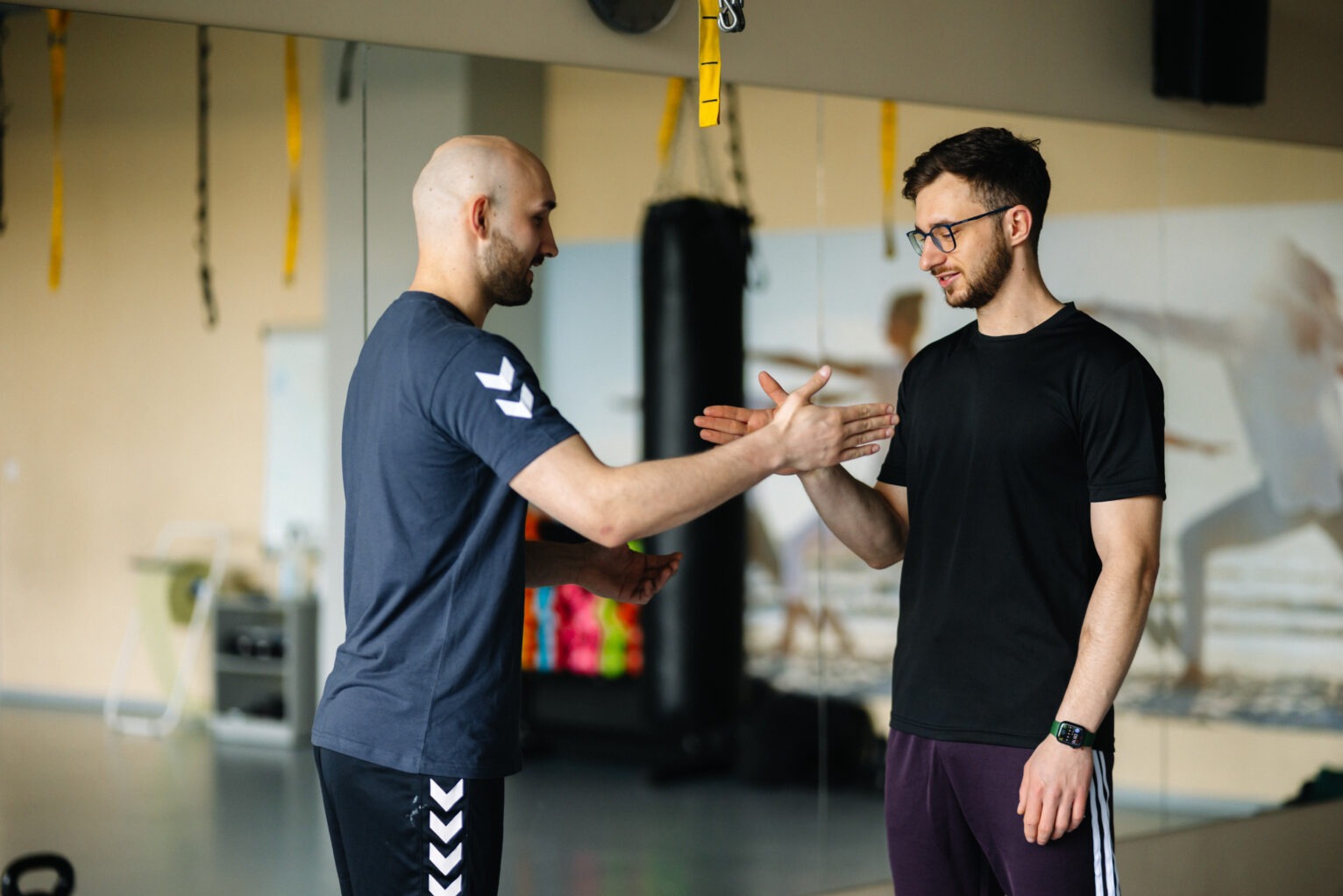 Dwóch mężczyzn w strojach sportowych wymienia się uściskiem dłoni w przestronnej siłowni z lustrami, widoczne worki treningowe i sprzęt do ćwiczeń.