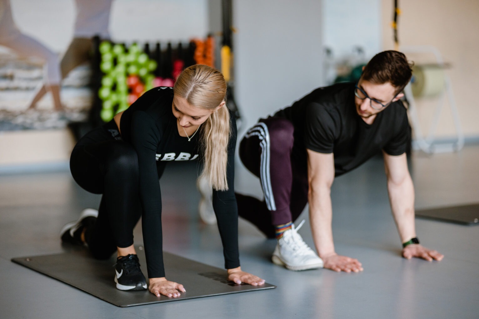 Kobieta z blond włosami i mężczyzna w okularach wykonują ćwiczenie rozciągające na macie w jasnym pomieszczeniu, ona w czarnym stroju sportowym, on w czarnej koszulce i spodniach dresowych...