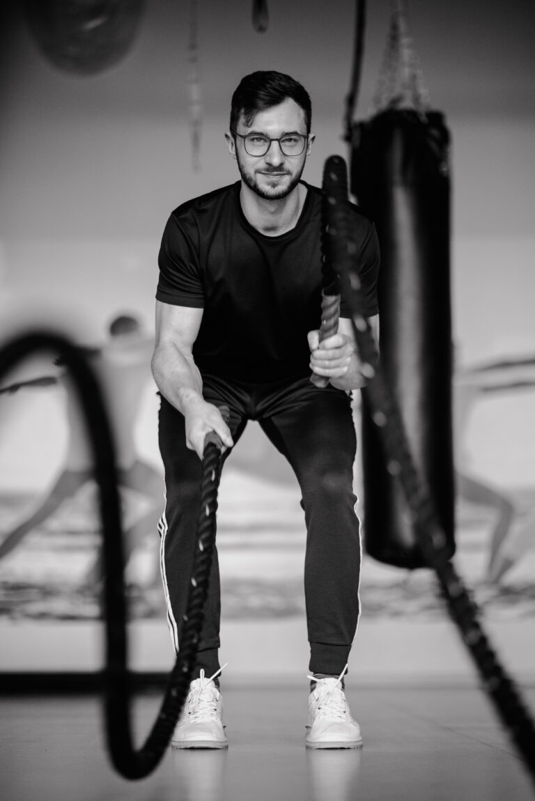 Czarno-białe zdjęcie mężczyzny w okularach, w sportowym stroju, trzymającego grube liny do ćwiczeń w siłowni, z workiem treningowym w tle.