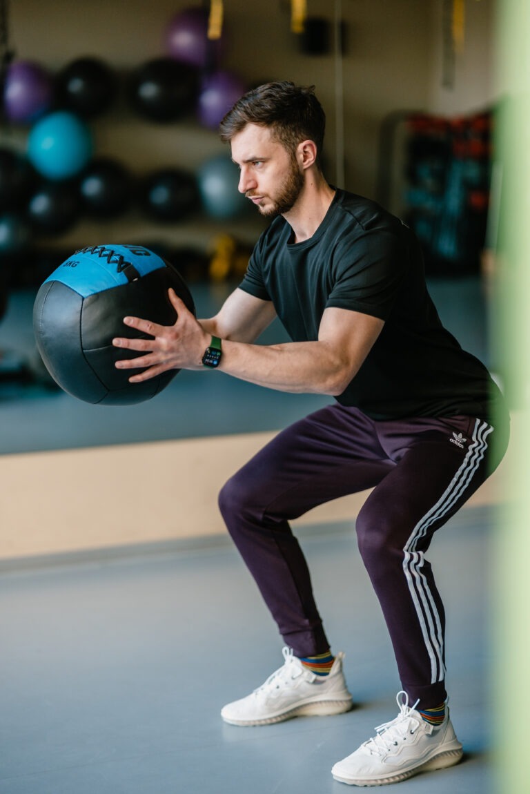 Młody mężczyzna w czarnej koszulce i fioletowych spodniach Adidas wykonuje przysiad z niebiesko-czarną piłką lekarską w siłowni, patrząc w prawo.