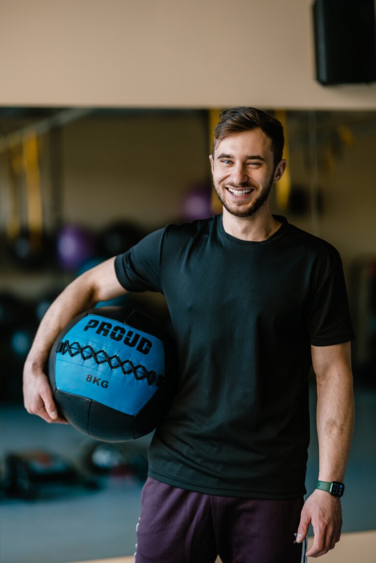 Uśmiechnięty trener personalny w czarnej koszulce trzyma niebiesko-czarną piłkę lekarską z napisem 'PROUD 8KG' w siłowni.