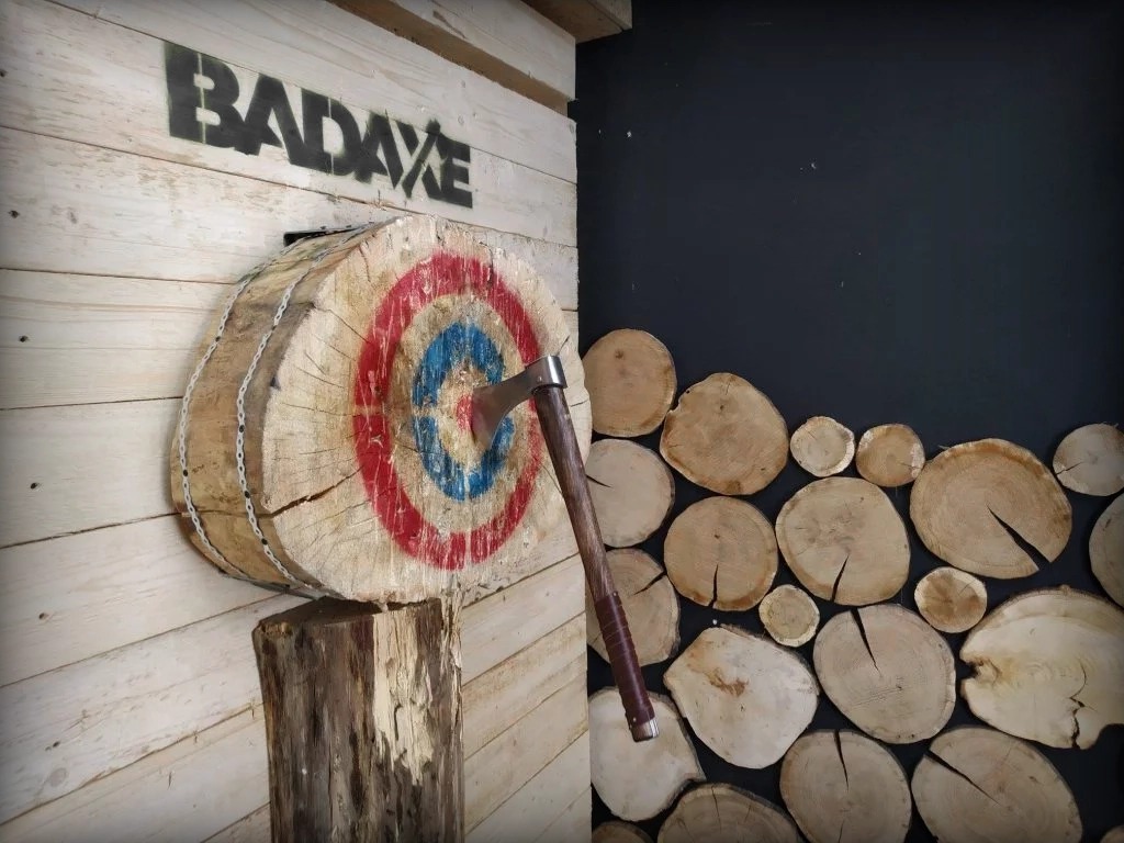 Topór wbity w drewnianą tarczę z namalowanym celem, zawieszoną na ścianie z jasnych desek z czarnym napisem 'BADAXE', obok ściana z ciemną farbą i dekoracją z drewnianych krążków.