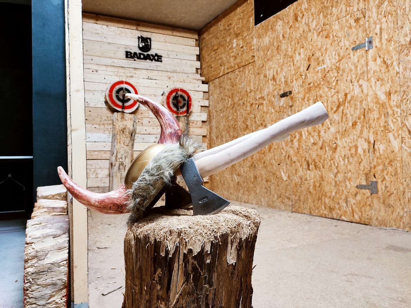 Topór i hełm wikinga z rogami na drewnianym pniu w hali do rzucania toporami, w tle tarcze do celowania.