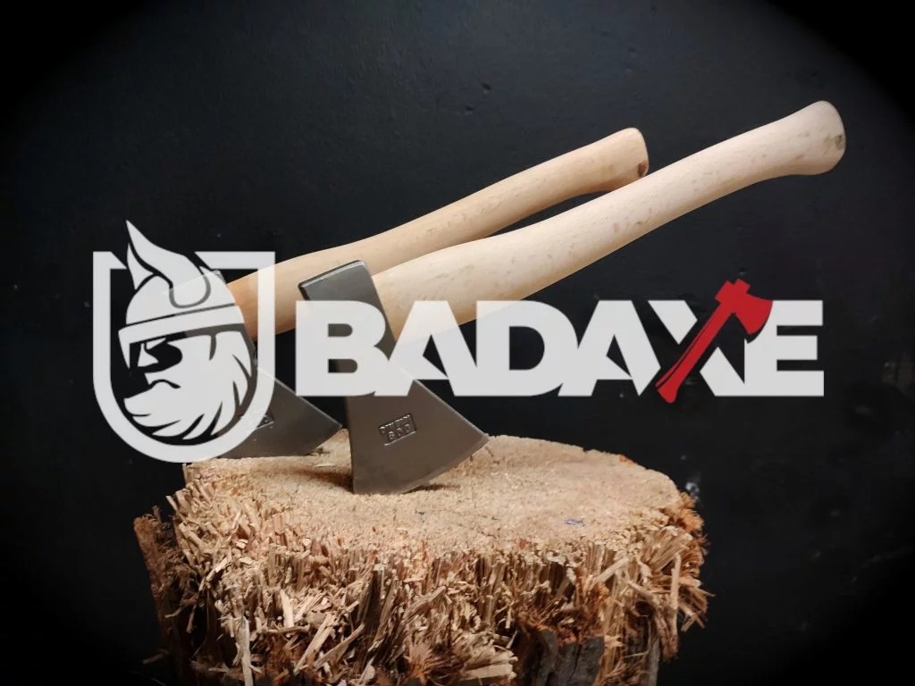 Dwa topory z jasnymi drewnianymi trzonkami, ułożone na pniu z wiórami, na tle logo firmy BADAXE z wizerunkiem Wikinga.