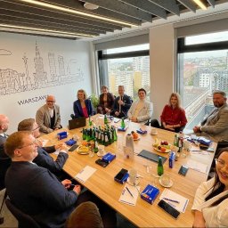 Alicja Małecka- Obiektywne doradztwo - Grupa osób w sali konferencyjnej podczas spotkania biznesowego, na ścianie grafika z panoramą Warszawy, na stole woda, owoce i materiały firmowe.