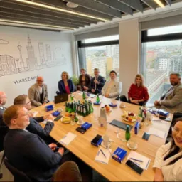 Grupa osób w sali konferencyjnej podczas spotkania biznesowego, na ścianie grafika z panoramą Warszawy, na stole woda, owoce i materiały firmowe.