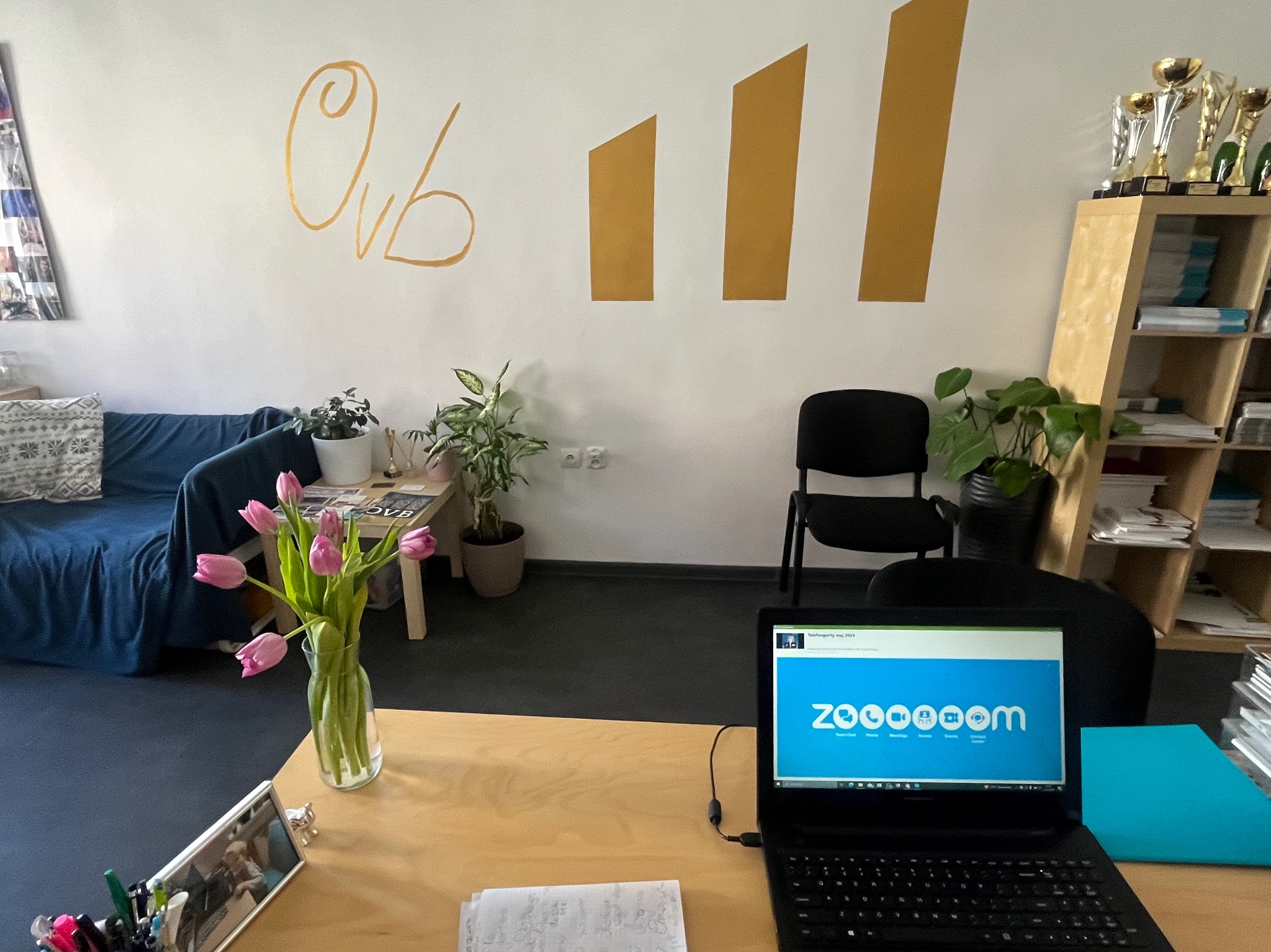 Biuro z biurkiem, laptopem z otwartą stroną Zoom, wazonem z tulipanami, sofą, regałem z trofeami i złotymi dekoracjami na ścianie.