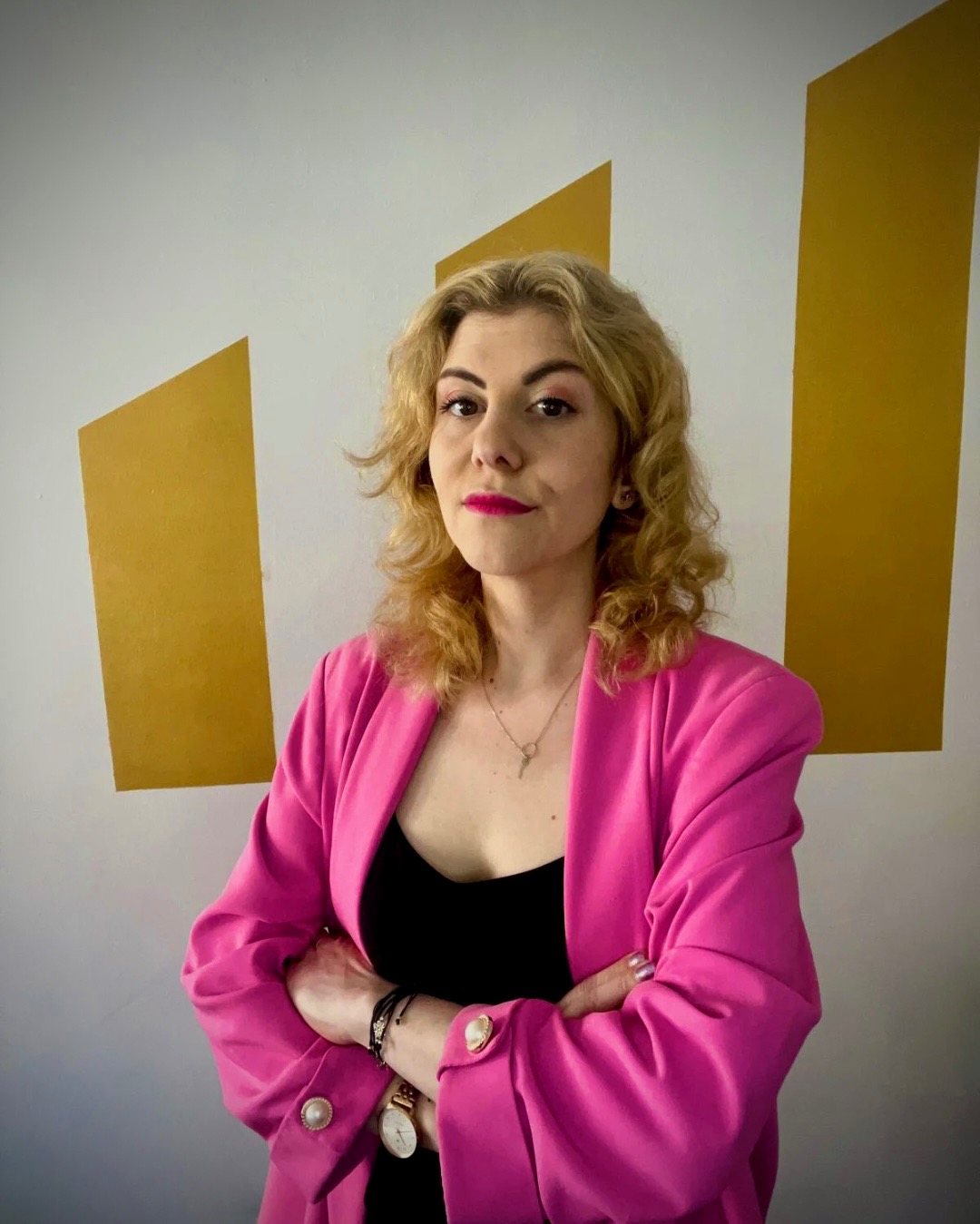 Portret kobiety w różowej marynarce z założonymi rękami na tle białej ściany z geometrycznymi złotymi elementami.