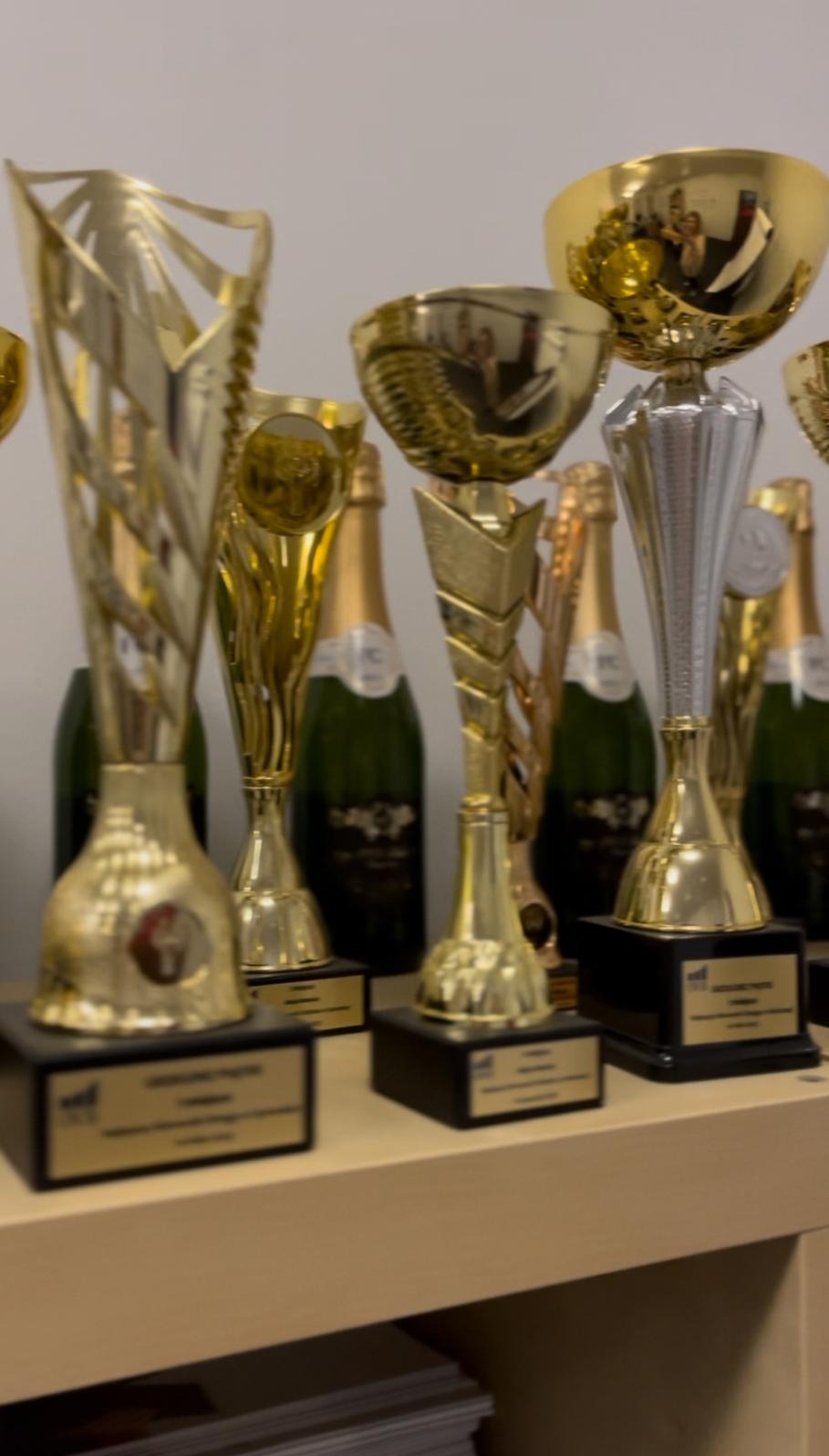 Półka z wieloma złotymi i srebrnymi pucharami oraz butelkami szampana w tle, symbolizująca sukces i osiągnięcia.