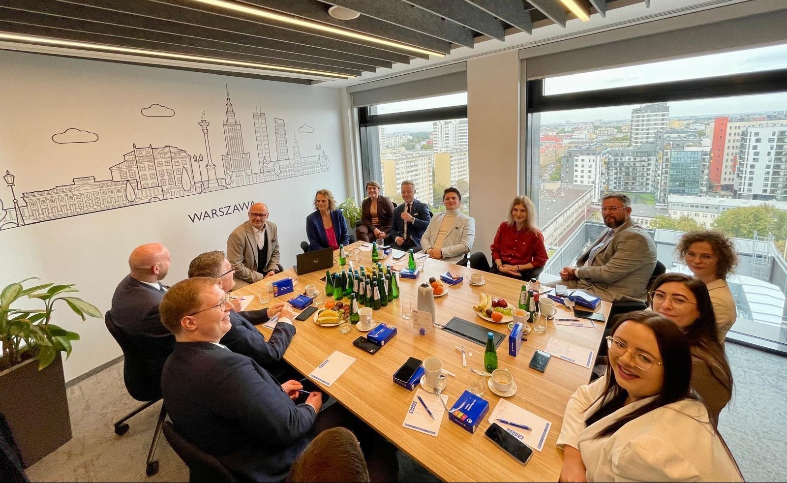 Grupa osób w sali konferencyjnej podczas spotkania biznesowego, na ścianie grafika z panoramą Warszawy, na stole woda, owoce i materiały firmowe.