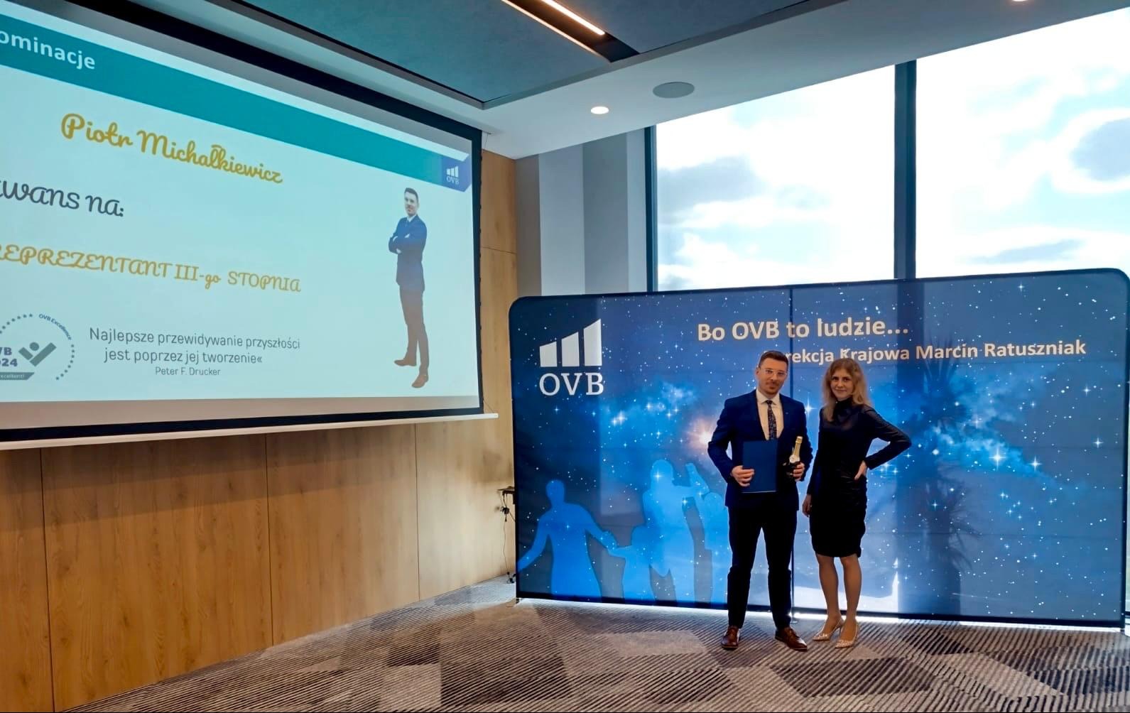 Wręczenie nagród OVB: mężczyzna w garniturze z dyplomem i butelką szampana, kobieta w czarnej sukience, tło z logo OVB i hasłem 'Bo OVB to ludzie...'