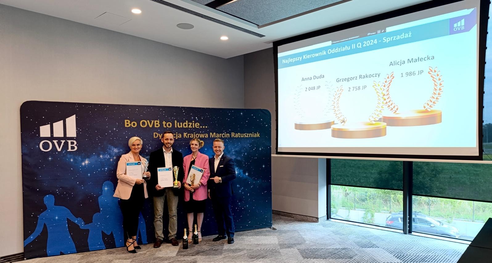 Cztery osoby, mężczyźni w garniturach i kobiety w żakietach, stoją na tle baneru OVB z logo i prezentacji wyświetlanej na ekranie, trzymając dyplomy i statuetki, prawdopodobnie podczas wręczenia...
