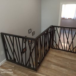 sPaweł Paweł Kurpierz - Metalowa balustrada schodowa z ukośnymi prętami i bramką zabezpieczającą na górze schodów, widoczne zdjęcia w ramkach na ścianie i fragment podłogi imitującej drewno.