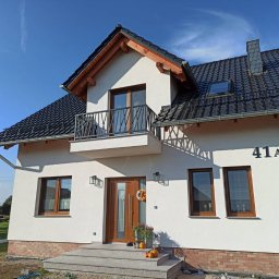 sPaweł Paweł Kurpierz - Nowoczesny dom jednorodzinny z balkonem z metalową balustradą o geometrycznym wzorze, brązowymi oknami i drzwiami, oraz czarnym dachem z dachówki ceramicznej.