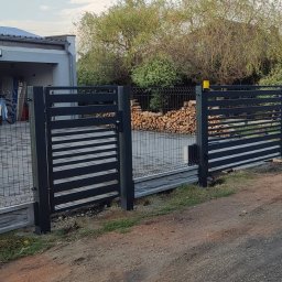 sPaweł Paweł Kurpierz - Nowoczesne ogrodzenie z paneli i bramka w kolorze antracytowym, z poziomymi szczeblami, na tle domu z garażem i stertą drewna.