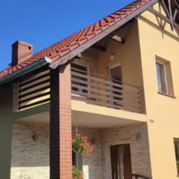 Dwukondygnacyjny dom z beżową elewacją, balkonem z poziomą balustradą i czerwonym dachem, otoczony zielenią.