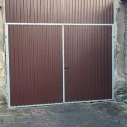 Brązowa, dwuskrzydłowa brama garażowa z metalowych paneli, z jasną ramą, zamontowana w starej, częściowo otynkowanej ścianie budynku z dachówką. Na dole widoczny podjazd z ażurowych płyt.