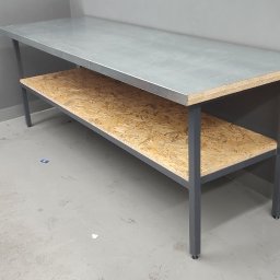 sPaweł Paweł Kurpierz - Metalowy stół warsztatowy z blatem ze stali nierdzewnej i półką z płyty OSB, ustawiony w rogu szarego garażu, obok narzędzi i pojemników.