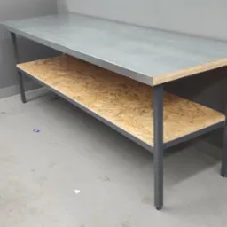 Metalowy stół warsztatowy z blatem ze stali nierdzewnej i półką z płyty OSB, ustawiony w rogu szarego garażu, obok narzędzi i pojemników.