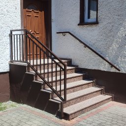 sPaweł Paweł Kurpierz - Zewnętrzne schody wejściowe z brązowego granitu z metalową balustradą w kolorze brązowym, prowadzące do drewnianych drzwi w białym budynku z oknem.