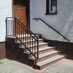 Zewnętrzne schody wejściowe z brązowego granitu z metalową balustradą w kolorze brązowym, prowadzące do drewnianych drzwi w białym budynku z oknem.