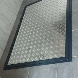 sPaweł Paweł Kurpierz - Metalowa klapa rewizyjna z blachy ryflowanej, wbudowana w podłogę z szarych płytek, widoczna z góry pod kątem.