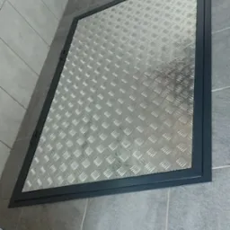 Metalowa klapa rewizyjna z blachy ryflowanej, wbudowana w podłogę z szarych płytek, widoczna z góry pod kątem.