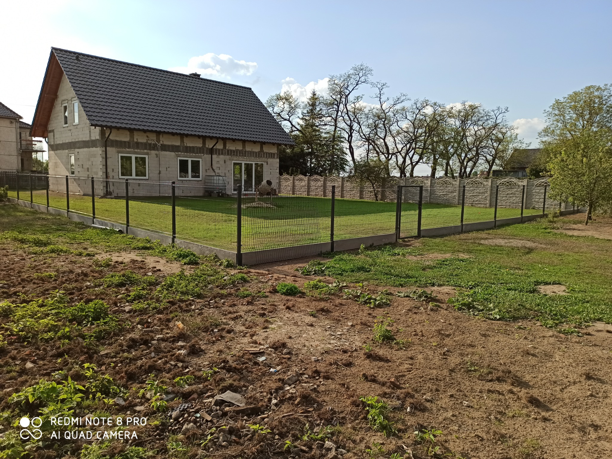 Nowoczesne ogrodzenie panelowe z metalowej siatki w kolorze grafitowym, zamontowane na betonowej podmurówce, otaczające trawnik przed domem w trakcie budowy. W tle widoczny fragment ogrodzenia...