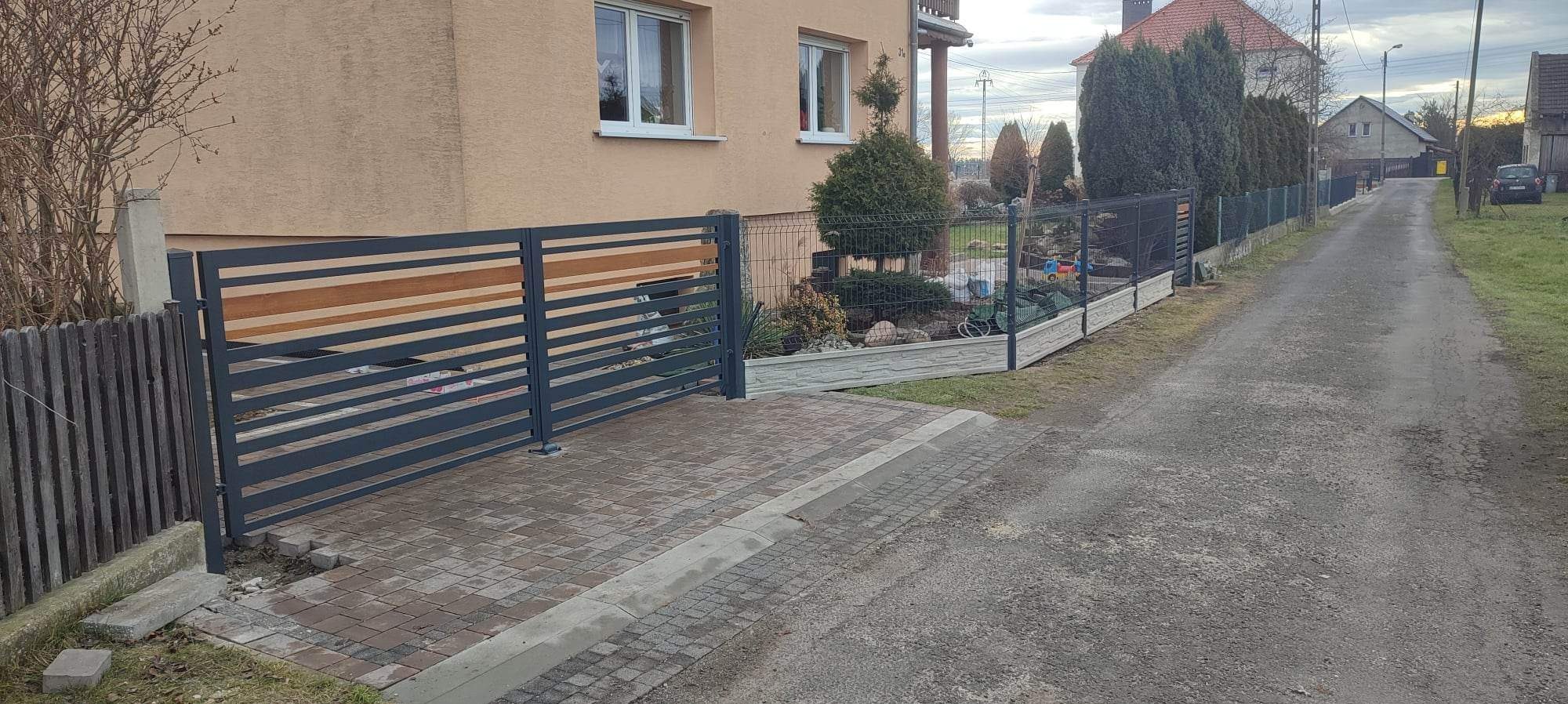 Nowoczesne ogrodzenie posesji z poziomych, grafitowych paneli z drewnianymi akcentami, przylegające do starszego, drewnianego ogrodzenia, kostka brukowa na podjeździe, widok na ulicę i domy w tle.