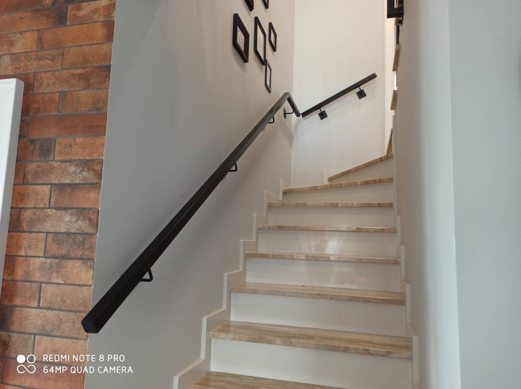 Wewnętrzne schody z marmurowymi stopniami i czarną metalową balustradą, zamontowaną na białej ścianie z dekoracją ceglaną w tle.