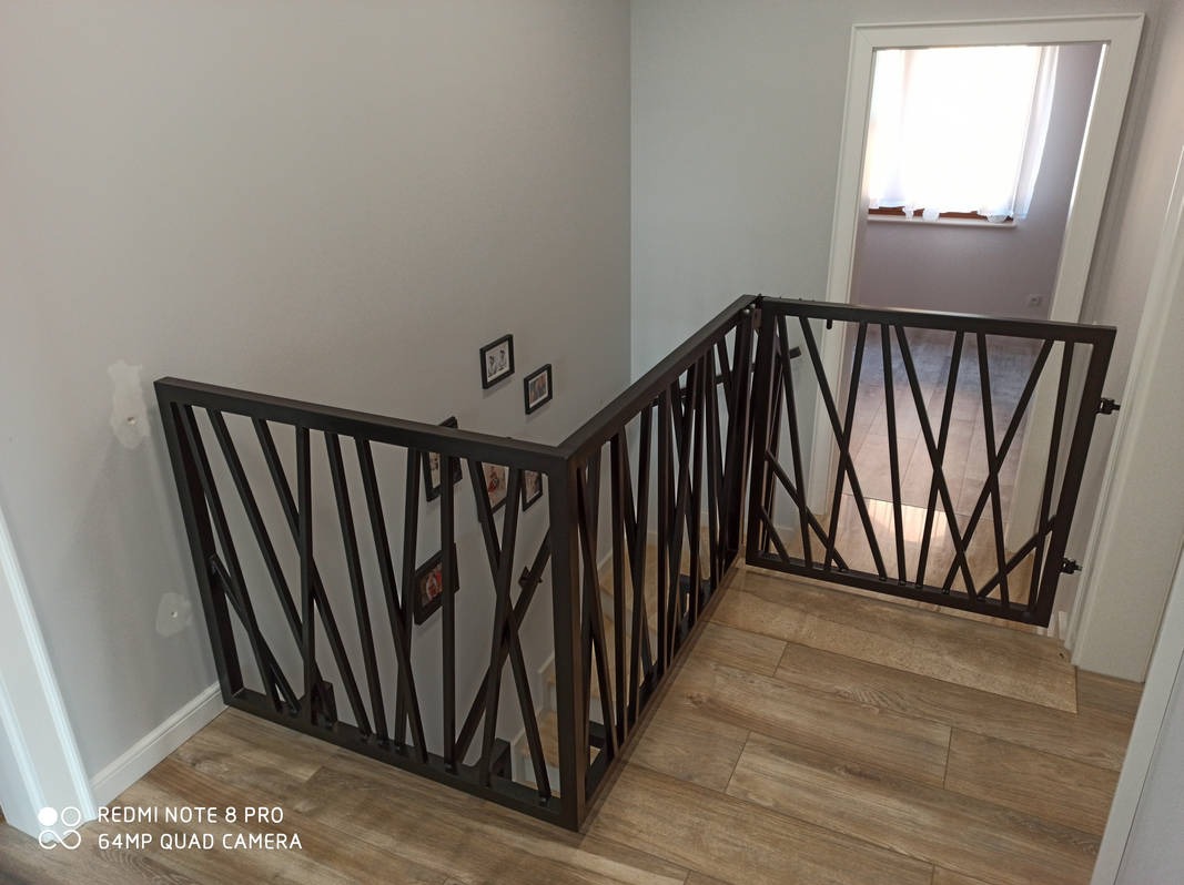 Metalowa balustrada schodowa z ukośnymi prętami i bramką zabezpieczającą na górze schodów, widoczne zdjęcia w ramkach na ścianie i fragment podłogi imitującej drewno.