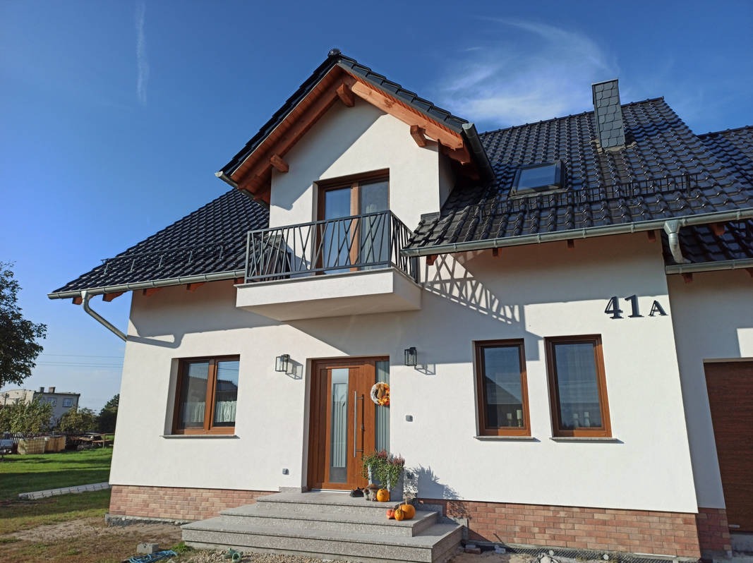 Nowoczesny dom jednorodzinny z balkonem z metalową balustradą o geometrycznym wzorze, brązowymi oknami i drzwiami, oraz czarnym dachem z dachówki ceramicznej.