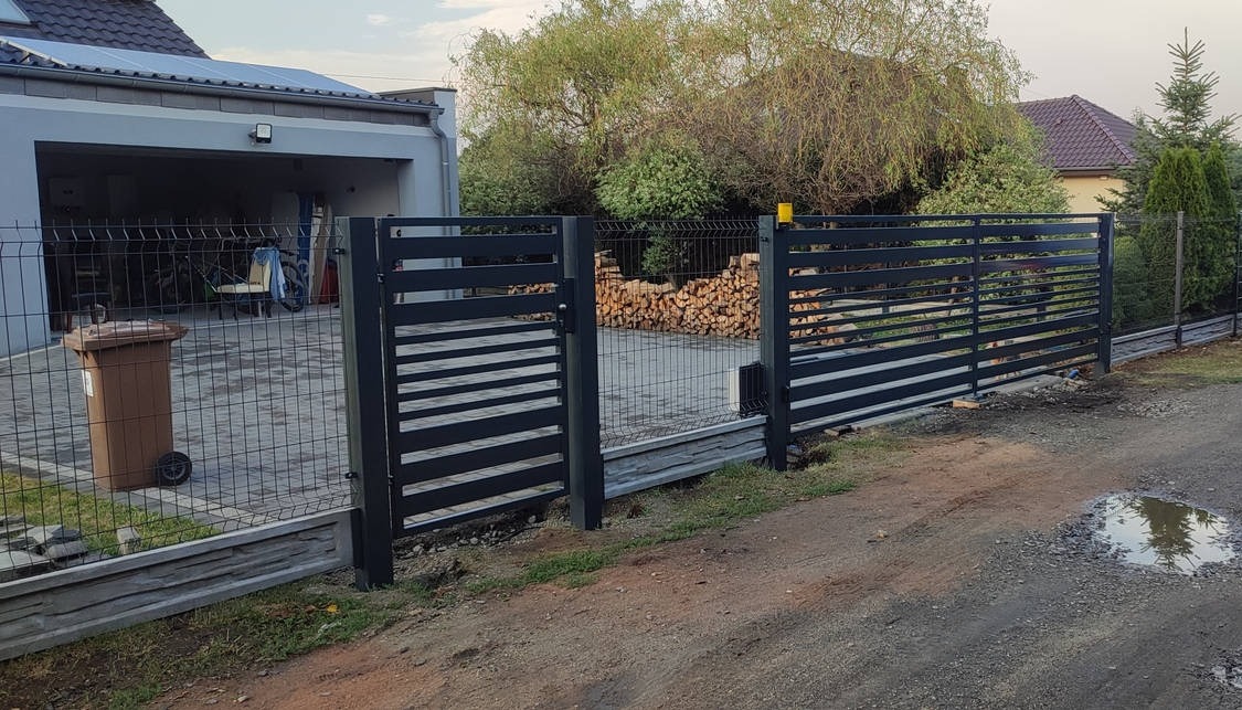 Nowoczesne ogrodzenie z paneli i bramka w kolorze antracytowym, z poziomymi szczeblami, na tle domu z garażem i stertą drewna.