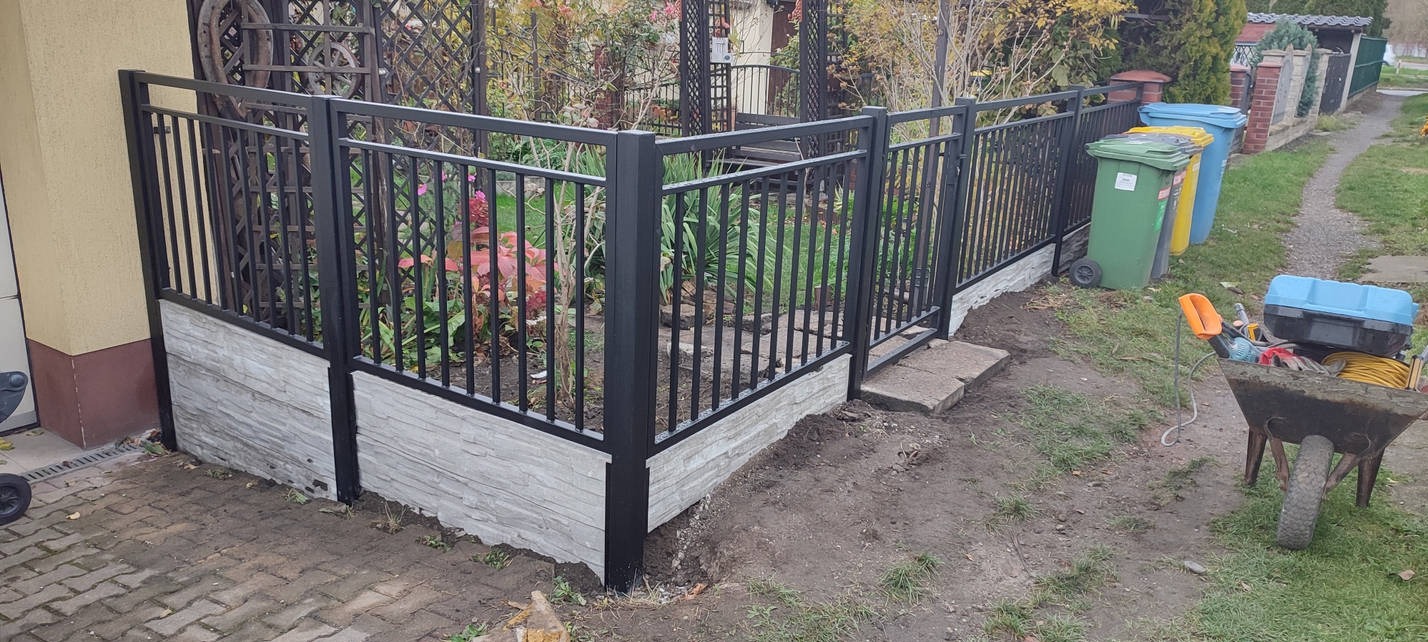 Częściowo ukończone, czarne ogrodzenie z metalowych prętów na niskiej podmurówce z szarej imitacji kamienia, widoczne ślady prac ziemnych i taczka z narzędziami obok.