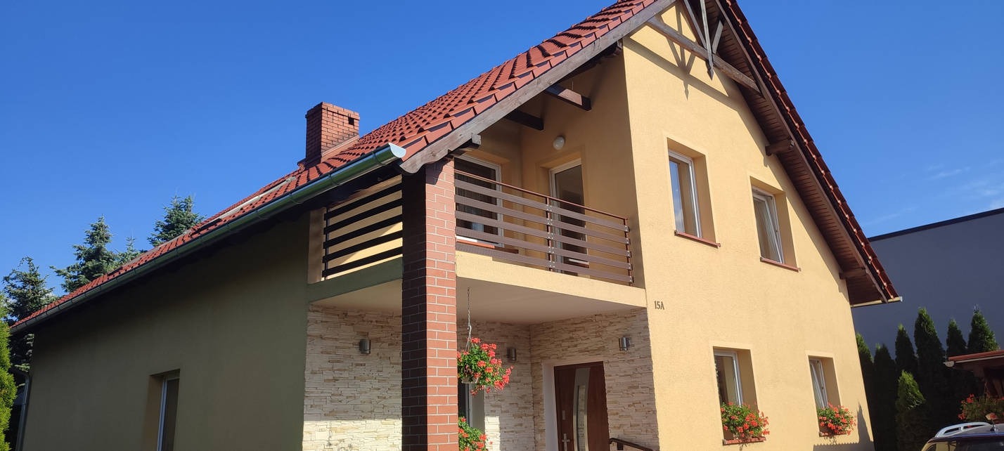 Dwukondygnacyjny dom z beżową elewacją, balkonem z poziomą balustradą i czerwonym dachem, otoczony zielenią.