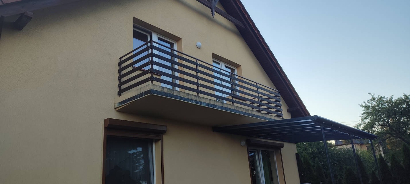 Nowoczesna balustrada balkonowa z poziomych, ciemnych profili, widoczna na tle elewacji domu oraz częściowo zadaszona pergola metalowa